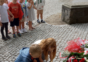 Dzieci stawiają zapalone znicze przed płytą, gdzie znajdują się wiązanki z biało-czerwonych kwiatów.