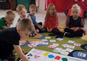 Chłopiec na macie do kodowania kładzie kartonik z niebieskimi kropkami, obok siedzą dzieci. W tle dekoracja z okazji Dnia Kropki.