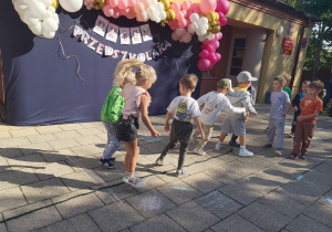 Przedszkolaki poruszają się tak, by nie dotknąć leżącej na ziemi liny, z tyłu dekoracja z balonów na niebieskim materiale.