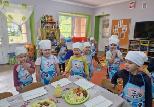 Przed nakrytym stołem stoją dzieci z zaciekawianiem spoglądając na półmiski z owocami.