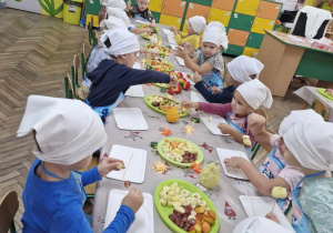 Dzieci siedzą przy stole i w skupieniu nadziewają owoce na patyczki.