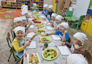 Siedzące wokół stołu przedszkolaki prezentują wykonane przez siebie owocowe przekąski.