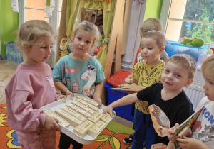 Dwie dziewczynki trzymają w rączkach tacę z upominkami dla chłopców: grami "domino".