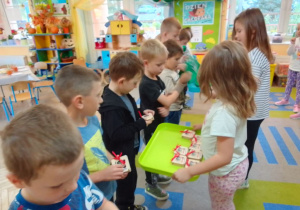 Chłopcy ustawieni w szeregu, przed nimi Wiktoria i Ala z tacami, na których są upominki dla chłopców: zielone torby i pudełka z grą.