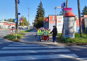 Dzieci ubrane w kamizelki odblaskowe stoją przed przejściem dla pieszych. Pani przypomina zasady bezpieczeństwa.