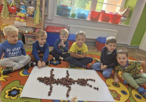 Karol, Ignaś, Olek, Franek, Szymon i Wojtek Z. siedzą na dywanie. Przed chłopcami na białym kartonie widać ułożoną z kasztanów mrówkę.