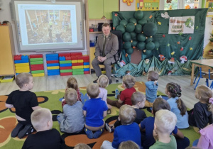 Pan leśnik siedzi obok tablicy interaktywnej, na której wyświetlana jest prezentacja multimedialna. Przed leśnikiem na dywanie siedzą dzieci.