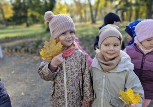 Ola i Alicja pozują do zdjęcia. Dziewczynki trzymają w dłoniach bukiety z jesiennych liści.