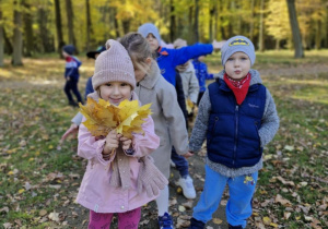 Dzieci idą alejką w parku. Na pierwszym planie widać Zuzię, która trzyma bukiet z liści.