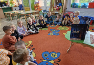 Dzieci siedzą w półkolu na dywanie, przed nimi na stoliku widać rozłożony teatrzyk.