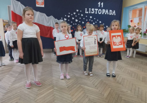 Czworo dzieci stoi na środku sceny podczas występu. Troje dzieci trzyma obrazki: z flagą, hymnem i godłem Polski.