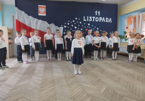 Dziewczynka stoi na środku sceny i deklamuje wiersz, a za nią stoją rówieśnicy. W tle dekoracja.