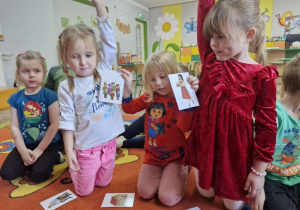 Nadia trzyma w rączkach dwa kartonika, obok Zuzia i Weronika trzymają dłonie w górze.