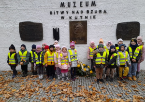 Dzieci stoją przed tablicami pamiątkowymi umieszczonymi na "Murze pamięci". Na murze znajduje się napis - Muzeum Bitwy nad Bzurą w Kutnie.