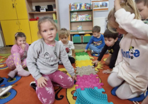 Dzieci siedzą na dywanie, na którym rozłożona jest ścieżka sensoryczna. Przedszkolaki dłońmi badają strukturę ścieżki.