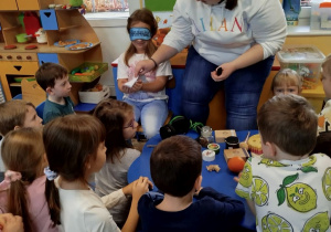 Lena siedzi na krzesełku z opaską na oczach, a Pani Martynka pokazuje, że możemy również woleć dotyk delikatny (chusteczka) lub ciężki (zabawka żaba wypełniona piaskiem).