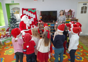 Dzieci stoją na dywanie odwrócone w kierunku Mikołaja, który siedzi na fotelu na tle dekoracji mikołajkowej. Obok stoi Pani dyrektor. Przedszkolaki mówią wiersz o Mikołaju.