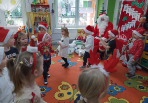 "Motylki" ustawione w kole naśladują jazdę samochodem i śpiewają piosenką "Jedzie Mikołaj". Występ dzieci ogląda Mikołaj, który siedzi na fotelu na tle dekoracji mikołajkowej.