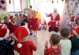 Przedszkolaki ustawione w półkolu przed Mikołajem śpiewają piosenkę.