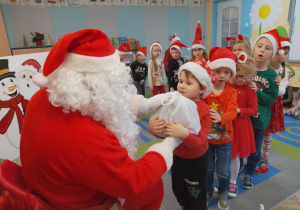 Dzieci w świątecznych strojach ustawione przed Mikołajem, Filip odbiera od Mikołaja upominek.