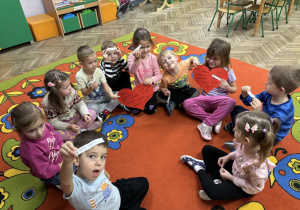 Dzieci siedzą w kole na dywanie i trzymają w rączkach andrzejkowe wróżby.