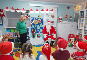 Mikołaj na tle świątecznej dekoracji siedzi na przygotowanym dla niego krześle. Obok Pani Grażynka pokazuje Mikołajowi listy narysowane przez dzieci. Dzieci ustawione są przodem do Mikołaja.