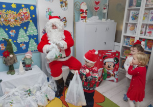 Chłopiec odbiera prezent od Mikołaja. 2 dziewczynki czekają na swoją kolej. Mikołaj siedzi na krześle wśród prezentów.