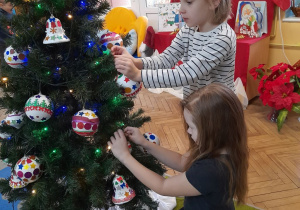 Dwie dziewczynki zawieszają swoje bombki na choince. W tle dekoracja świąteczna.