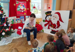 Mama Uli czyta dzieciom książkę "Świąteczne opowieści. Misia i jej mali pacjenci" autortwa A. Cholewińskiej-Szkolik. W tle choinka z bombkami, świąteczna dekoracja (plakat Mikołaja i bałwanka) oraz książki opowiadające o świętach.