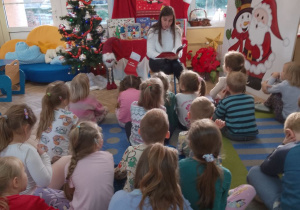 Przedszkolaki siedzą na dywanie. Mama Julii czyta dzieciom kolejną opowieść z książki "Święty Mikołaj wchodzi kominem" pt. "Święty Mikołaj pociesza smoka". W tle dekoracja świąteczna.