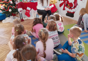 Mama Julii siedzi na krześle, a przed nią na dywanie dzieci. Zaproszony gość rozmawia z dziećmi na temat wysłuchanej opowieści. W tle dekoracja świąteczna.