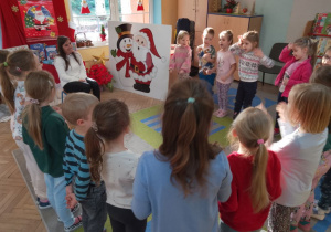 Mama Julii siedzi na krześle, a przed nią stoją dzieci i śpiewają piosenkę o Mikołaju.