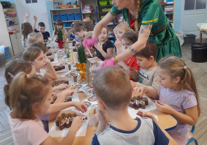 Dzieci siedzą przy złączonych stołach i ozdabiają swoje czekoladowe choinki. Pani rozdaje przedszkolakom kolejne dodatki.