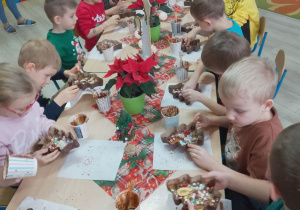 Dzieci siedzą przy stoliczkach z ozdobionymi choinkami.