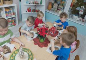 Druga część grupy przy stoliku wrzuca czekoladowe kulki z formy do kubków, w tle świąteczna dekoracja sali.