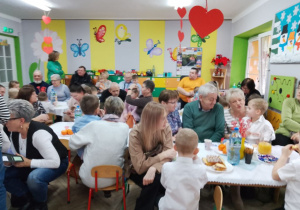 Przedszkolaki wraz ze swoimi dziadkami siedzą przy stolikach, na których widać smakowite słodkości.