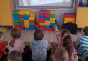 Dzieci siedzą na dywanie przed tablicą interaktywną i oglądają film edukacyjny o walentynkach.