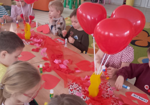 Dzieci siedzą przy wspólnym stole i wykonują kartki walentynkowe w kształcie serc.