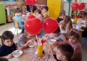 Dzieci siedzą przy stolikach i spożywają posiłek. Na stolikach znajdują się wazoniki z czerwonymi balonami w kształcie serc oraz serwetki w serduszka.