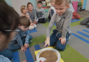 Dzieci uczestniczą w eksperymencie "Jak bakterie uciekają od mydła". Na dywanie stoi płaski talerz z wodą i mielonym pieprzem. Obok siedzą dzieci. Antek zanurzył palec w wodzie, a dzieci obserwują jak bakterie (pieprz) przykleiły się do palca.