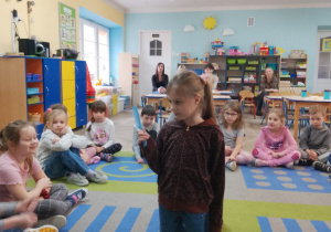 Dzieci siedzą na dywanie, a Zuzia przykucnęła na przed tacą z przyborami toaletowymi. Dziewczynka wybrała grzebień i określa jego przeznaczenie. Rodzice siedzą na krzesłach i obserwują działania dzieci.