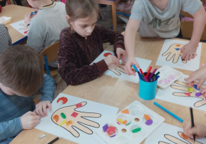 Dzieci przy stolikach naklejają na sylwetę ręki naklejki piankowe i dorysowują flamastrami elementy.