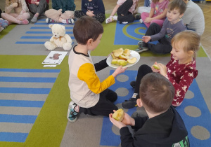 Przedszkolaki siedzą na dywanie, a Mikołaj częstuje dzieci jabłkami. Na środku dywanu siedzi Miś, a przed nim leży karta uważności "Miś je".