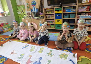 Dzieci siedzą na dywanie. Przed nimi na białym kartonie widać ułożone z części obrazki z wizerunkami dinozaurów.