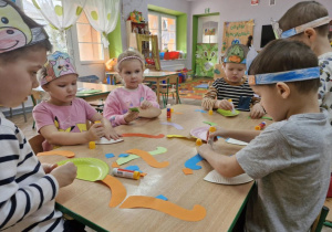 Dzieci z grupy Motylków siedzą przy stolikach w trakcie wykonywania pracy plastycznej. Przedszkolaki tworzą wizerunki dinozaura z papierowych talerzyków.