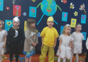 Na scenie Weronika przebrana za brudną Ziemię oraz Lucjan przebrany za Księżyc i Franek przebrany za Słońce. Obok widać dziewczynki w strojach gwiazdek.