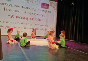 Dzieci siedzą na scenie, pomiędzy nimi leży jasnozielona chusta.