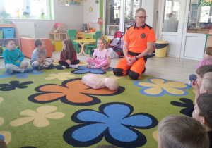 Dzieci siedzą w kole na dywanie obok nich siedzi ratownik medyczny. Na środku leży fantom.