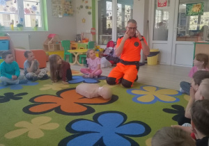 Dzieci siedzą w kole na dywanie obok nich siedzi ratownik medyczny, który wskazuje na swoje oczy, utrwala w ten sposób numer alarmowy 112.