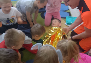 Pan Krzysztof razem z dziećmi okrywa folią termiczną Mikołaja, który leży na dywanie.